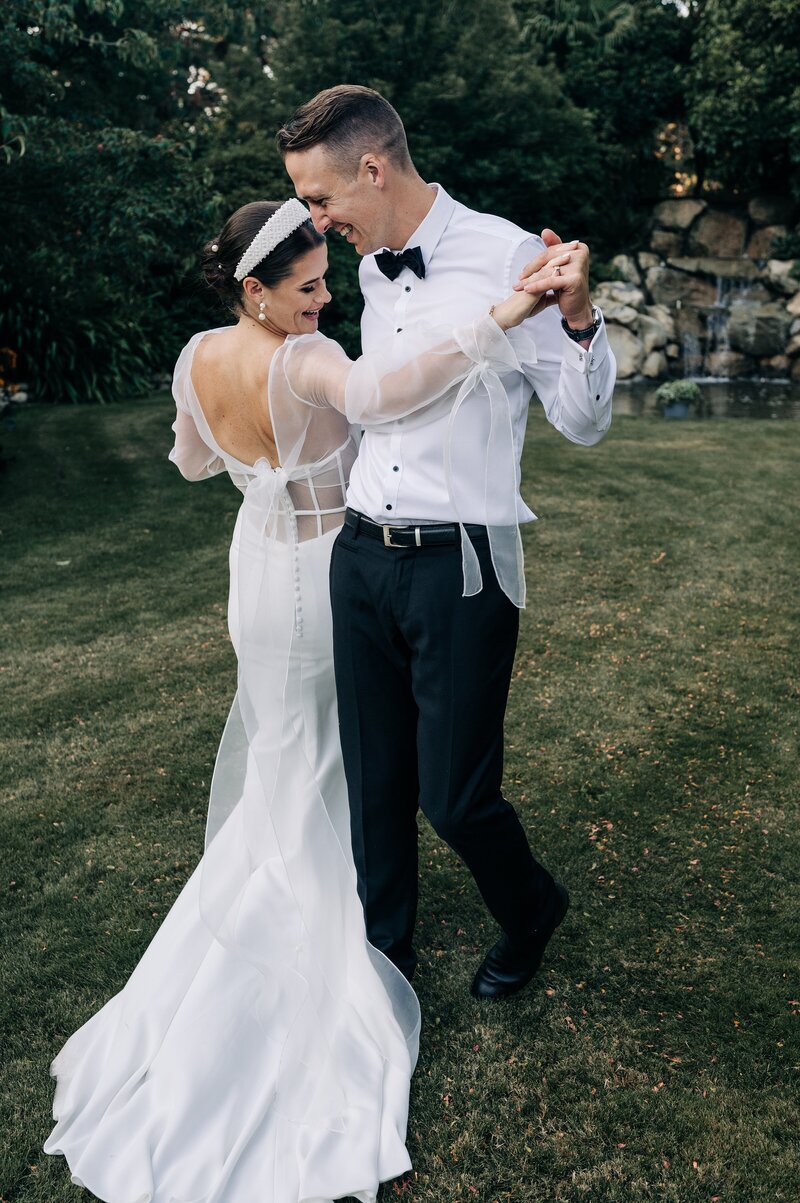 bride in trish peng gown and groom in black bowtie dance on the lawn at sunset at their christchurch wedding