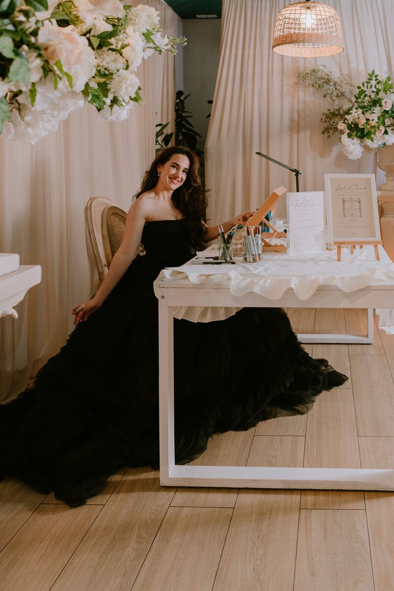 A woman in a strapless black gown sits at a white table with art supplies, smiling while creating live illustrations at an event, surrounded by elegant floral arrangements and soft draped curtains.