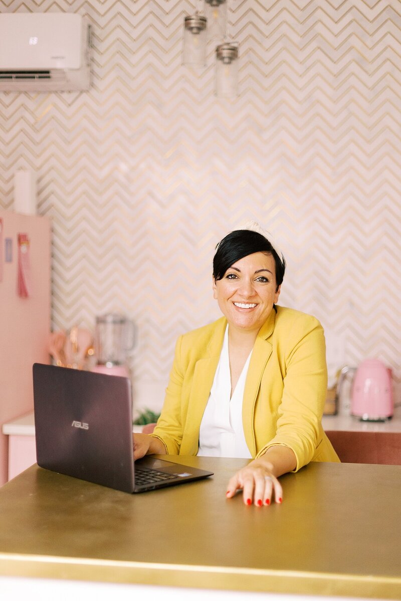 A woman with a pixie hair cut is standing at a kitchen counter and working on a computer and is smiling up at the camera at the Nashville Pinky House