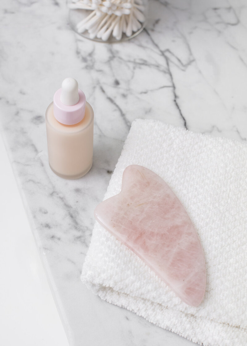pink gua sha stone with white towel and pink tincture bottle containing skincare on white marble countertop