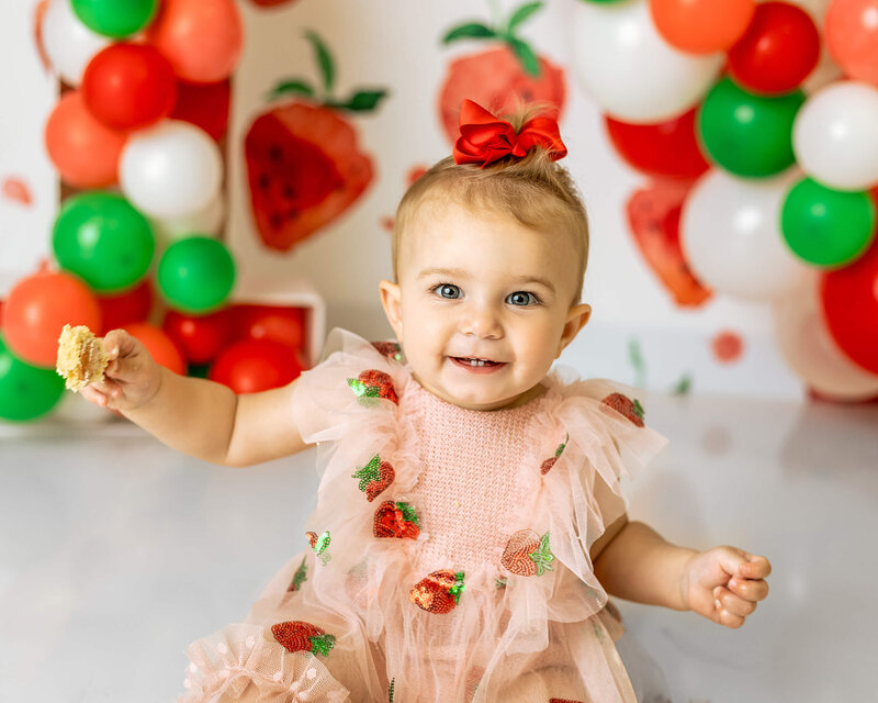 A little girl wearing a pink strawberry romper is holding a spoon with cake and smiling