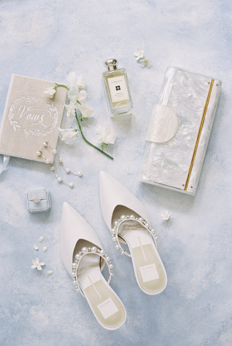 bridal shoes vow book and jewelry in a flat lay image with a cloud blue background for a classic Cape Cod wedding