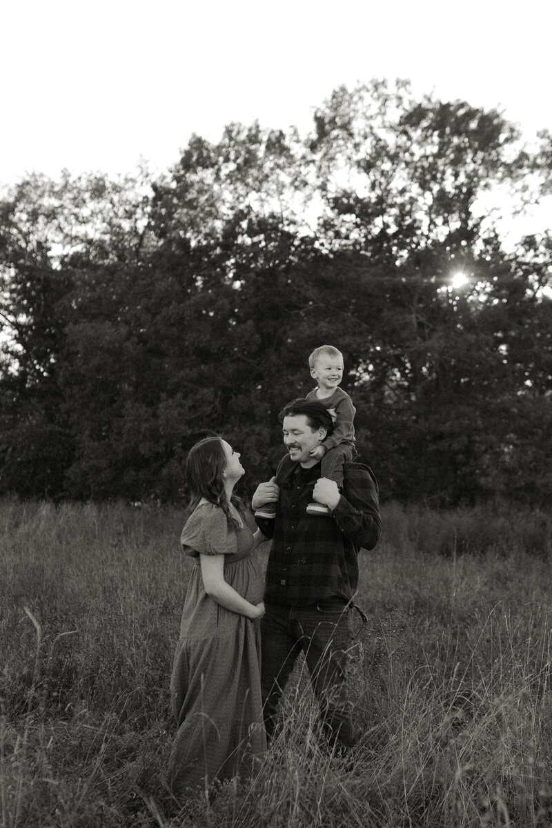 Mom and dad embrace summer family photos to announce a baby on the way, as their toddler perches on his dad's shoulders.