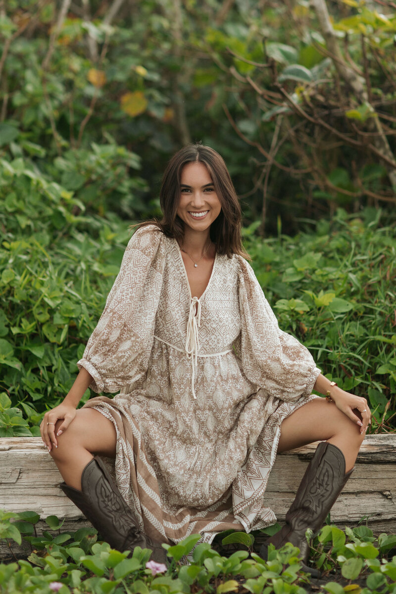 A woman wearing a flowy dress, smiling and sitting on a log in nature