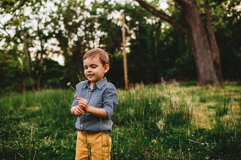 little boy in yellow pants and a denim shirt blowing a dandelion into the wind with the woods in the distance captured by Baltimore photographers