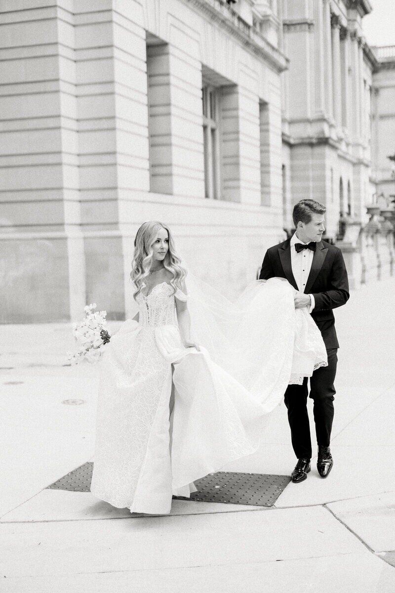 Groom gently holding the bride’s wedding dress train as they walk together outdoors, captured by a Milwaukee wedding photographer.