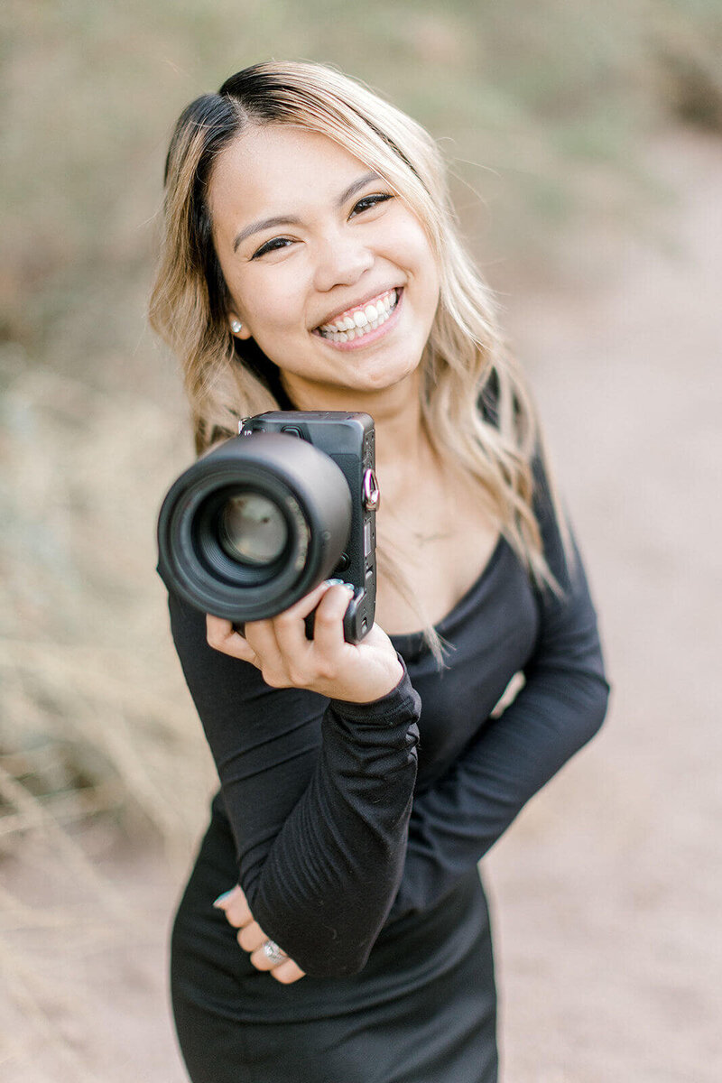 A portrait of a female photographer holding her camera in front of herself, smiling.