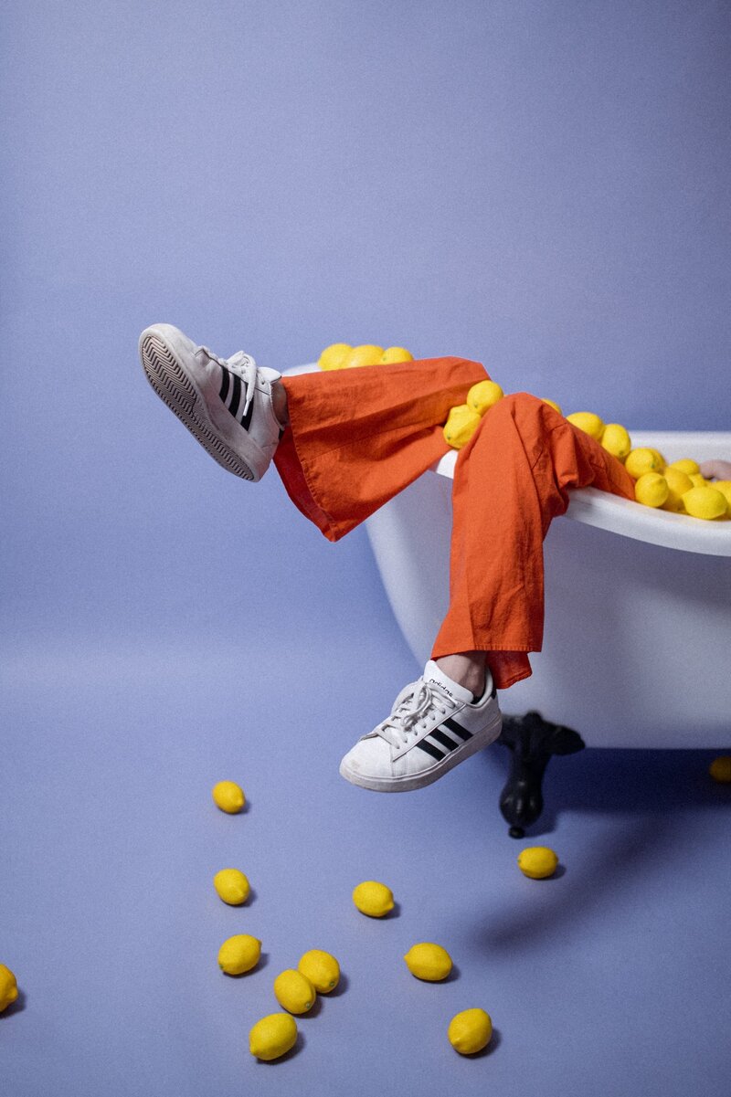 Legs lay over the edge of a bathtub wearing orange pants and adidas shoes. The bathtub is overflowing with lemons against a purple backdrop.