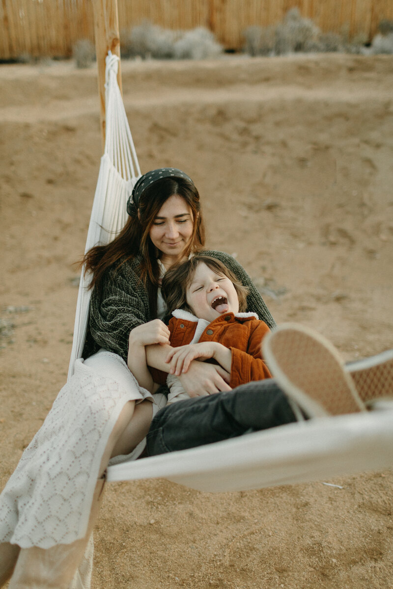 A mother and her young child share a joyful moment in a hammock, surrounded by a sandy landscape. the child is laughing heartily as the mother smiles gently, holding him close