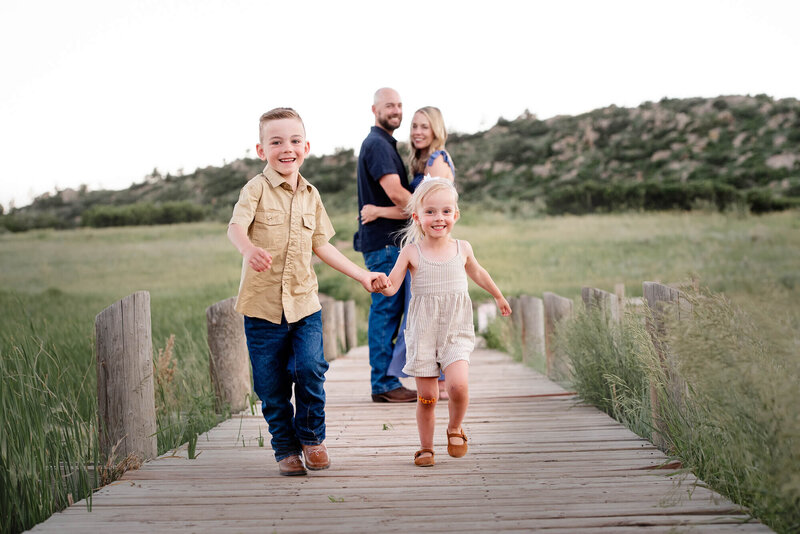 A toddler boy holds hands and runs with his little sister on a bridge from mom and dad