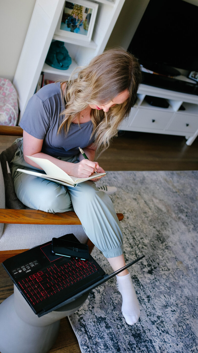 Sammy sitting with her legs over the arm of a chair writing in a notebook with her laptop on the table next to her