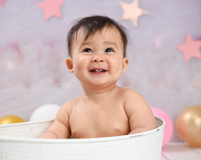 A baby sitting in a white tub with a pink and gold star-themed background, smiling and showing small teeth