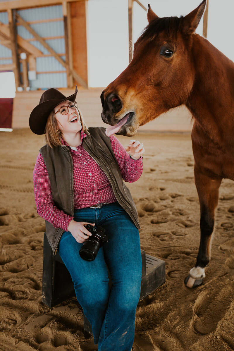 a cowgirl sitting on a stool with her camera looking up to her horse standing next to her
