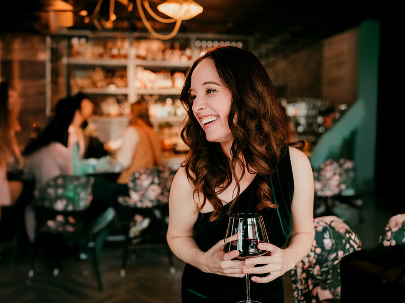A woman laughing with a glass of wine in a stylish bar, representing a confident and natural dating photo style—featured in a photography quiz to help users find their ideal dating profile look.
