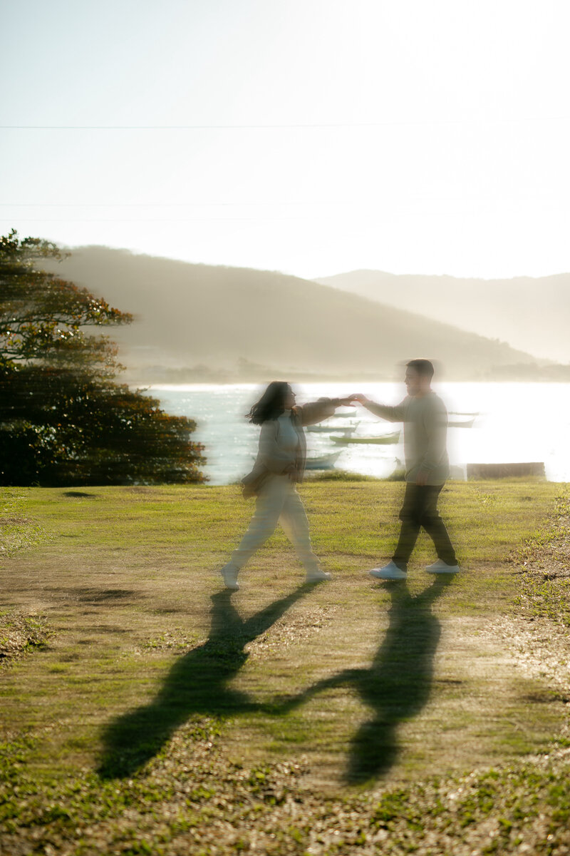 A couple dances on a sunlit grassy field with blurred motion, casting long shadows. The background features a serene lake and distant mountains.