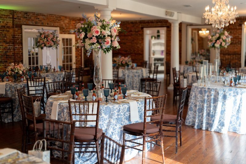 Elegant wedding reception setup at The White Room in historic downtown Saint Augustine featuring round tables with blue and white patterned linens, tall floral centerpieces with pink and white flowers, mahogany chiavari chairs, exposed brick walls, crystal chandeliers, and hardwood floors