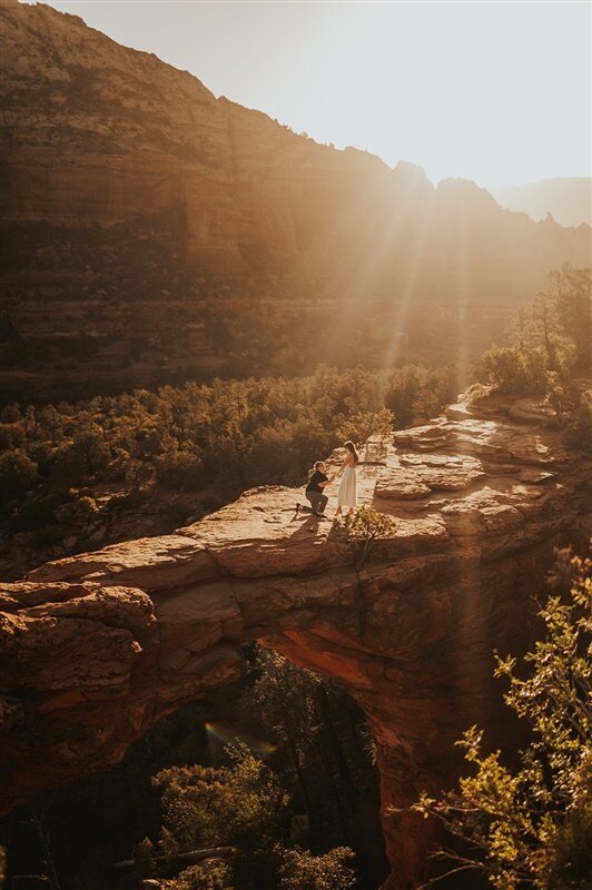 A couple stands on a dramatic rock formation during their proposal in Sedona, Arizona, with golden sunlight streaming down around them. The red rock landscape and distant mountains create a breathtaking backdrop.