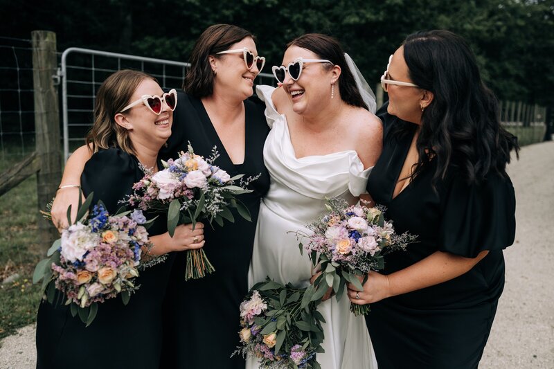 bridal party in black dresses with white heart sunglasses laugh together by deer fence at bangor farm