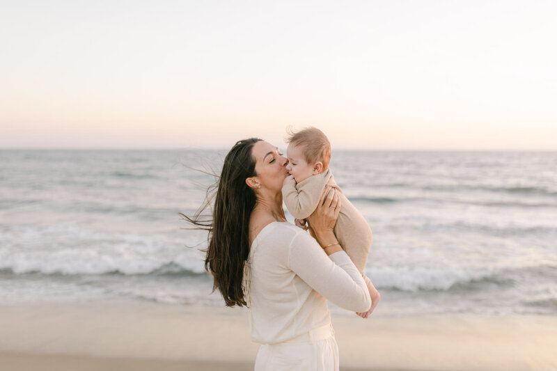 Orange County Family Photographer - mother holding her baby up on a beach while kissing his cheek