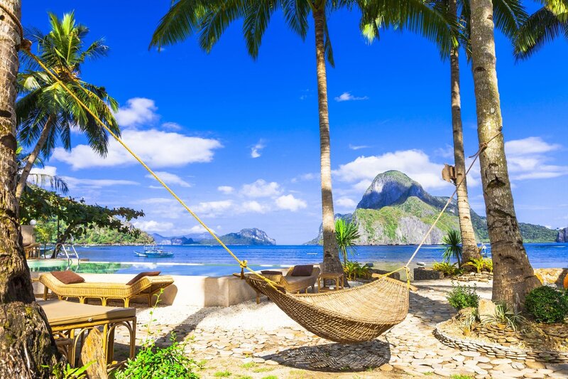 Tropical Bali resort with a hammock between palm trees, pool, ocean view, and rocky mountain in the distance.