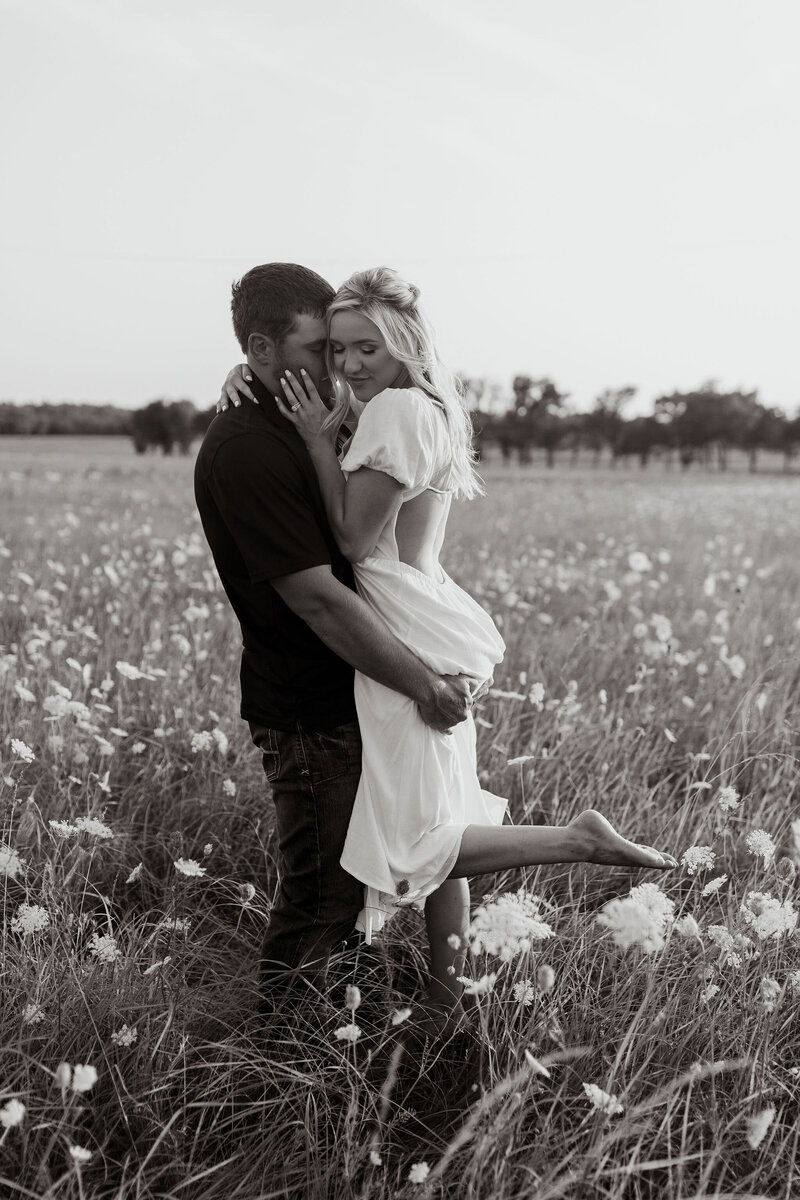 man lifting up woman in white dress in front of wildflower field