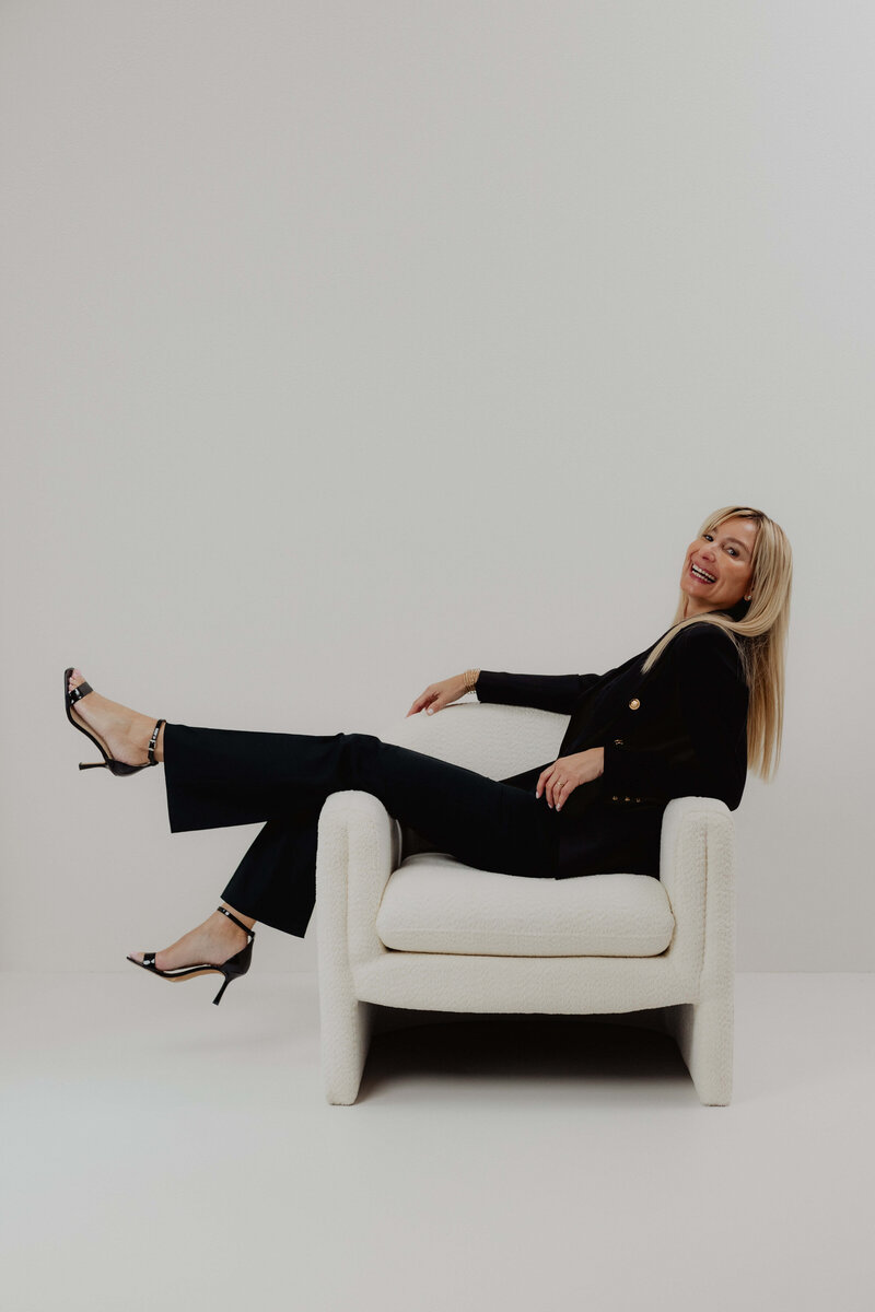 A women in a black power suite and heels swinging her legs and sitting in a white chair.
