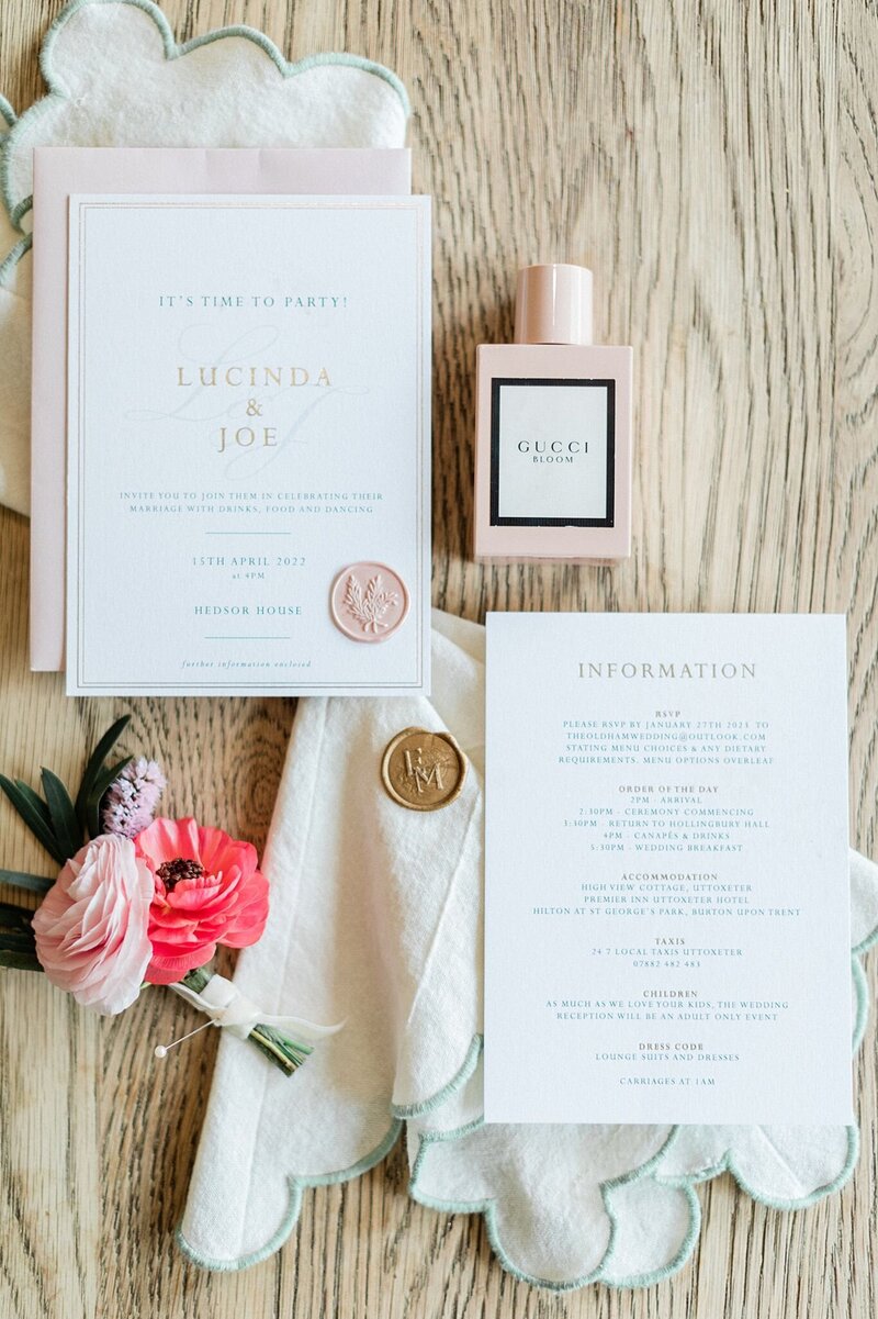 Elegant wedding stationery flatlay featuring blush and white invitation suite, Gucci Bloom perfume, wax seals, and a single ranunculus flower