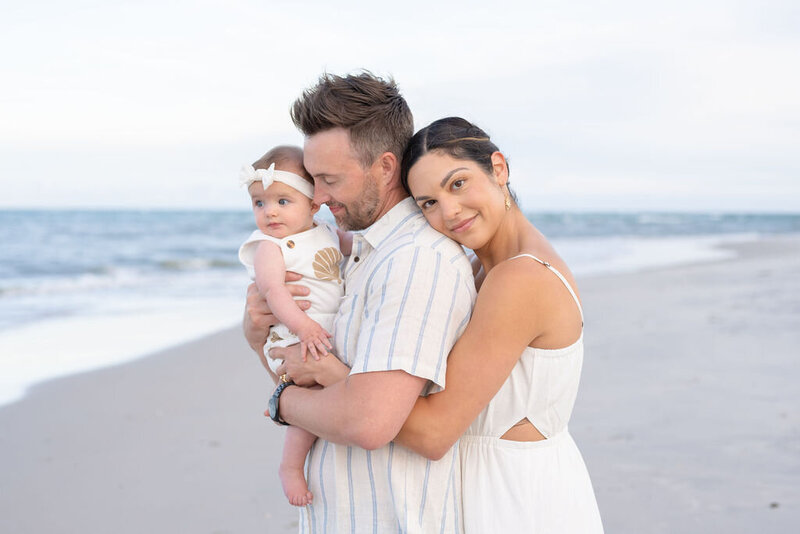 closeup of father holding a baby girl wearing a white bow on north topsail beach as mother hugs his back and rests her head with the tide is coming in and the sky is white and clear