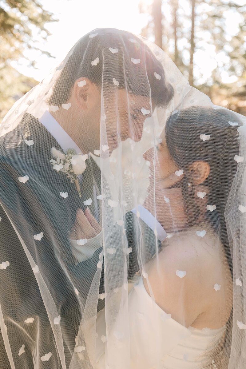 wedding couple snuggling and smiling under the brides veil in the sunlight at glacier point in yosemite during their elopement photoshoot