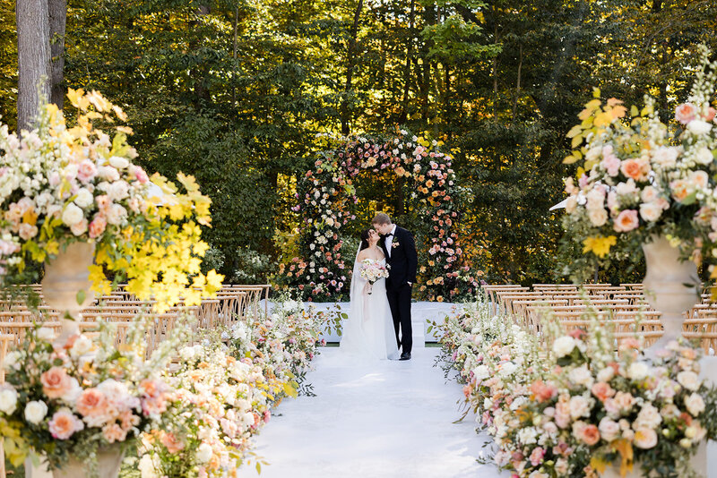 Luxurious late summer outdoor wedding venue with floral arch, and lush aisle flowers plus urns.   