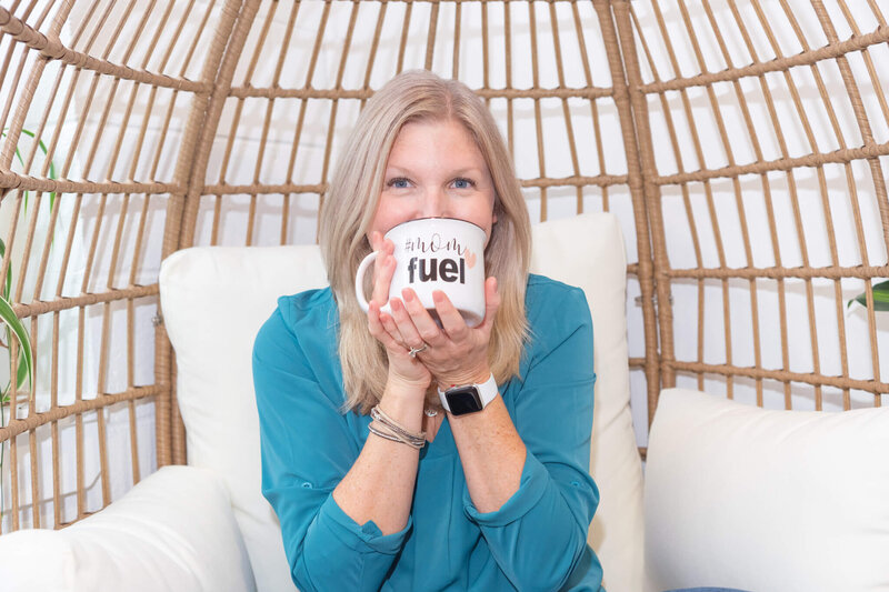 Maureen in teal shirt sitting in chair  drinking coffee from a mug that says Mom fuel