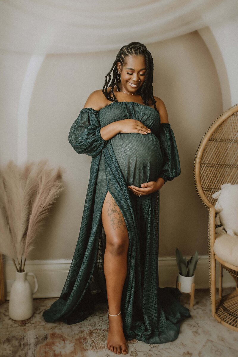 A pregnant woman in a green off-shoulder dress smiles while cradling her baby bump, captured beautifully by a maternity photographer in Perry, GA. She stands barefoot beside a wicker chair and a vase of pampas grass in a softly lit room.