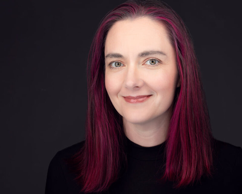 Alicia Insley Smith, Fort Mill Portrait Photographer white woman with pink highlights in her dark hair, green hazel eyes, smiling at the camera wearing black on a dark background, traditional headshot crop