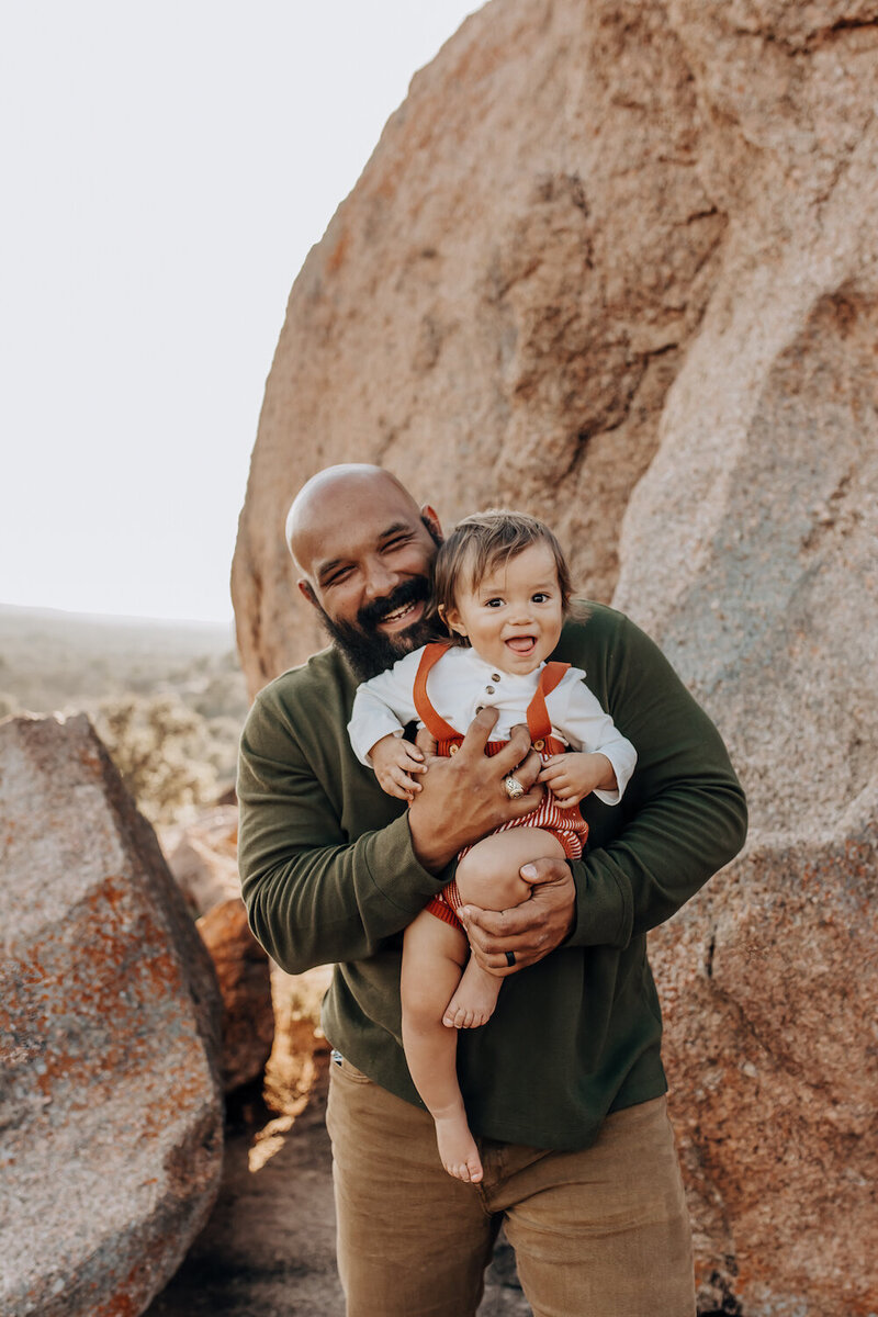 A dad smiles proudly while holding his son atEnchanted rock state natural area in Fredericksburg, Texas.