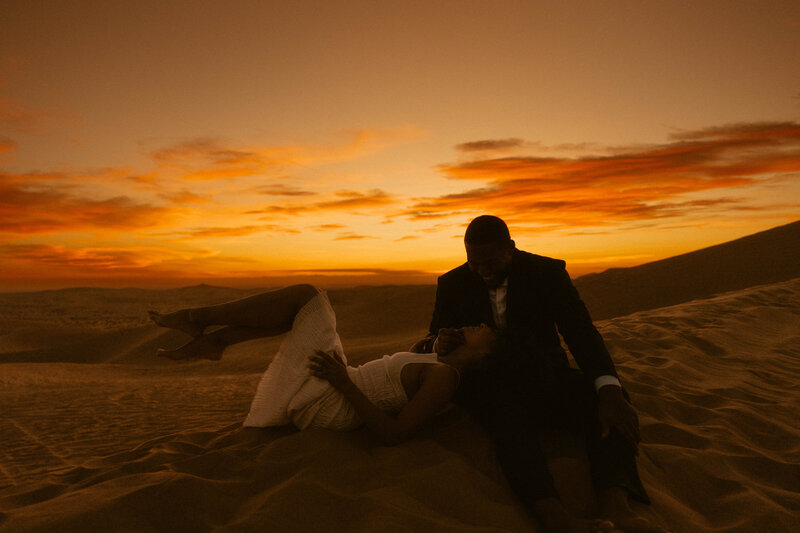 a man and woman laying in the sand during a sunset