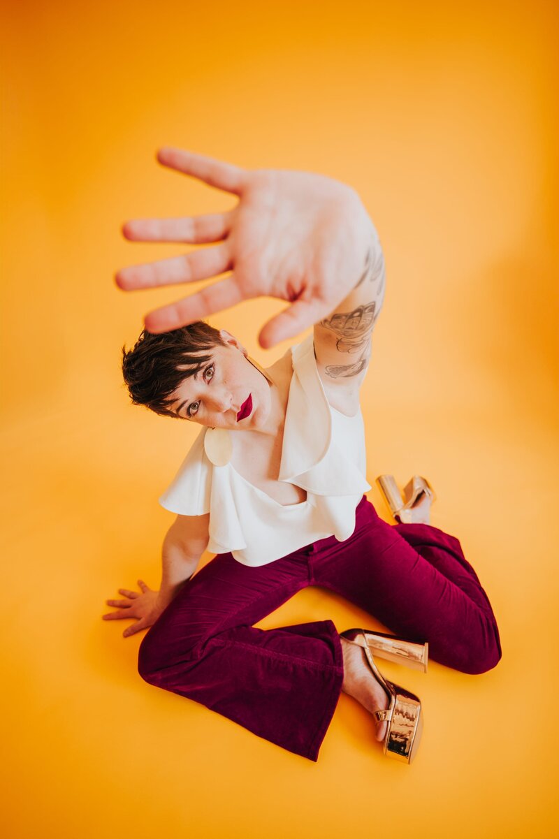 Woman with very short brown hair sits in a cream-colored flowy shirt , burgundy pants and heels with hand outstretched toward the camera in front of a yellow-orange backdrop.