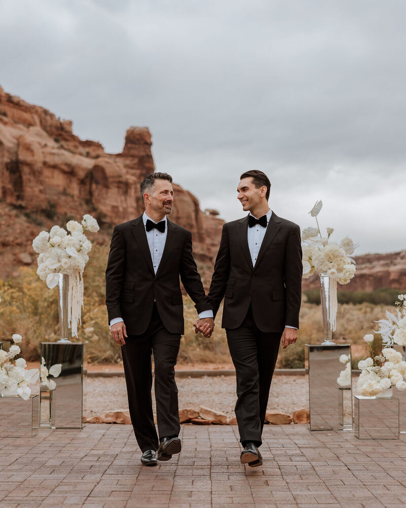 two grooms holding hands white walking down wedding aisle in the desert