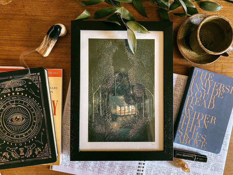 A framed picture of a den in the woods lays on a table amongst palo santo, journals and notebooks.