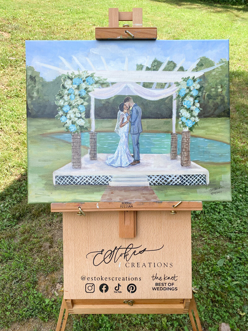 Easel displaying an acrylic painting of an interracial couple's first kiss at their outdoor wedding ceremony, highlighting an intimate and romantic moment