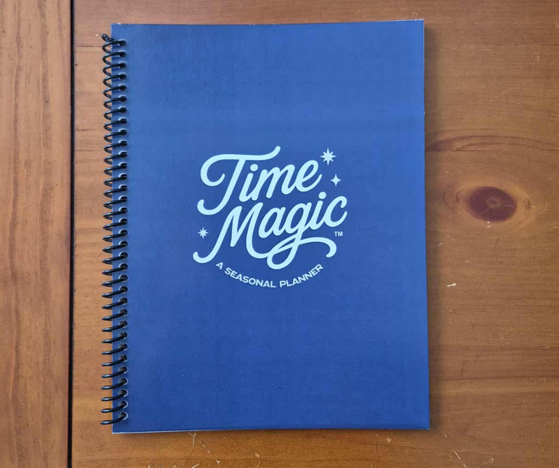 A navy blue spiral-bound Time Magic Seasonal Planner placed on a wooden surface, showing the cover with the white Time Magic logo.