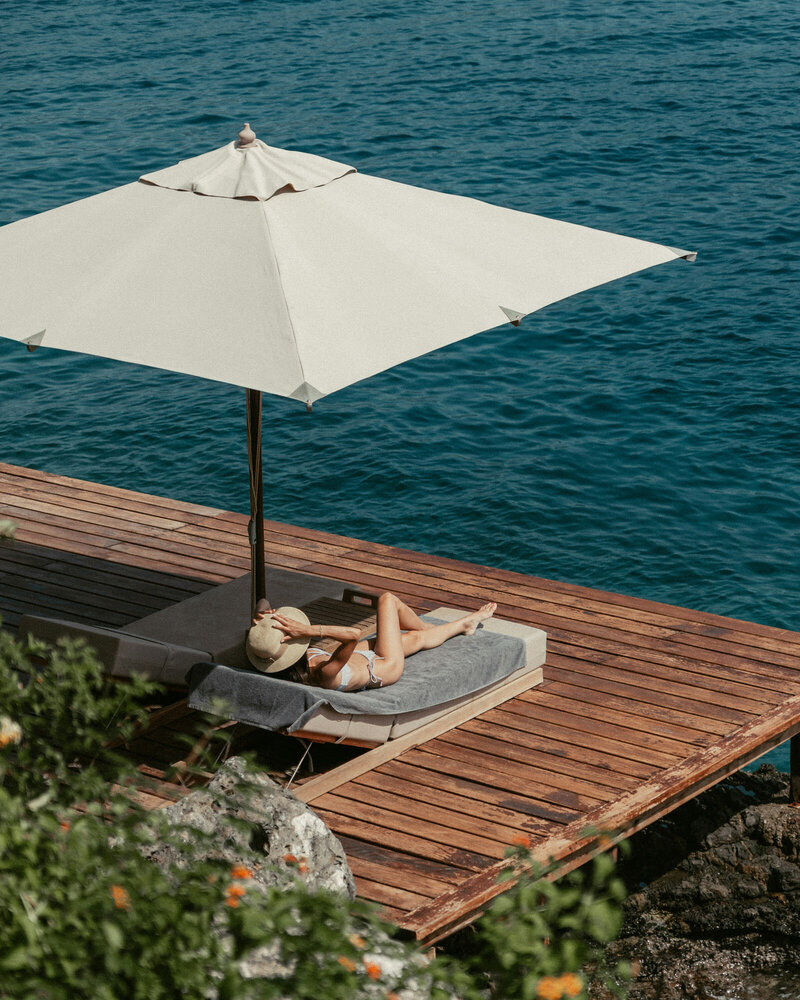 Woman laying on a lounge chair under an umbrella on a dock.