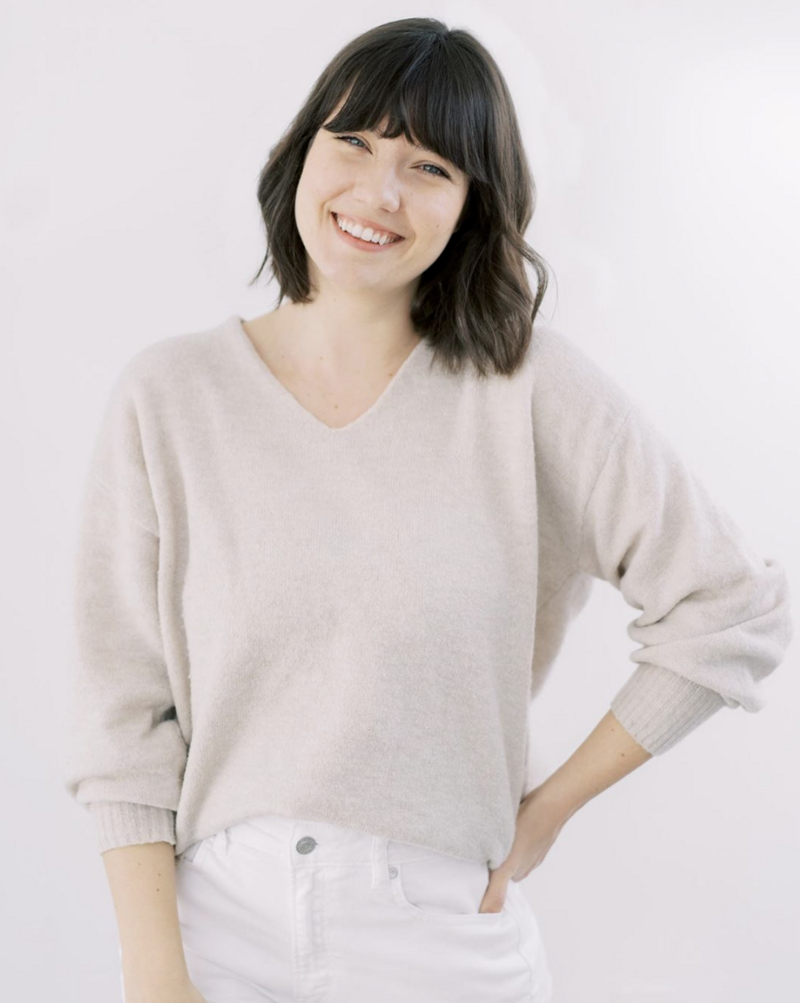 An image of Jess Imrie, smiling at the camera wearing white pants and cream sweater with her hand resting on her hip.