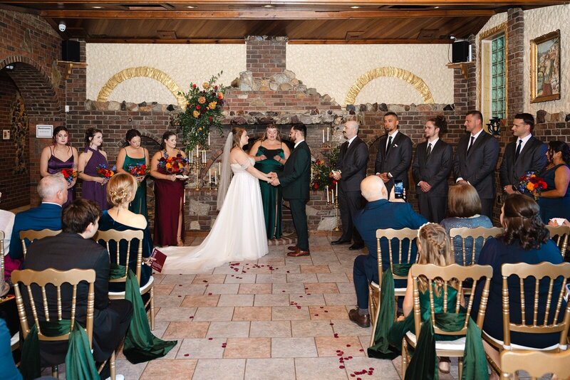 A bride and groom hold hands during their indoor wedding ceremony, surrounded by their wedding party and guests at Grbic Events in St. Louis.