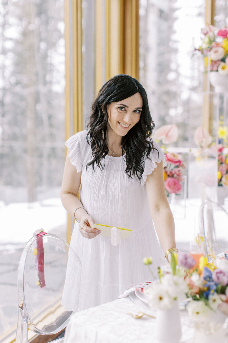 A woman with long dark hair wearing a white dress stands at a table set with flowers, holding a piece of paper. She is indoors with large windows in the background, meticulously attending to details as part of her role in partial wedding planning for a Banff wedding.