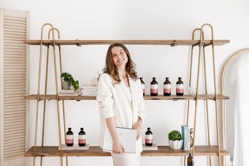 Smiling woman in cream blazer holding a notebook, standing in front of a shelf lined with naturopathic products