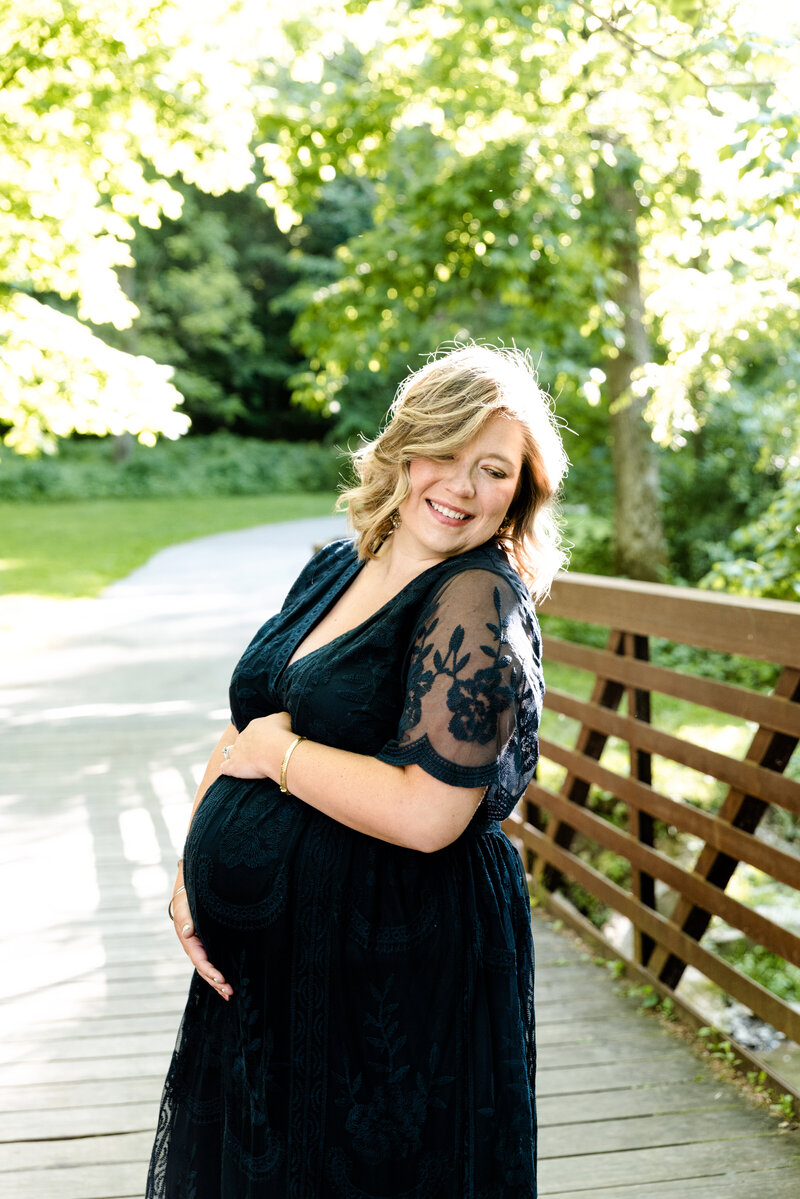 beautiful pregnant mom in long nacy lace dress holding belly and smiling on a bridge