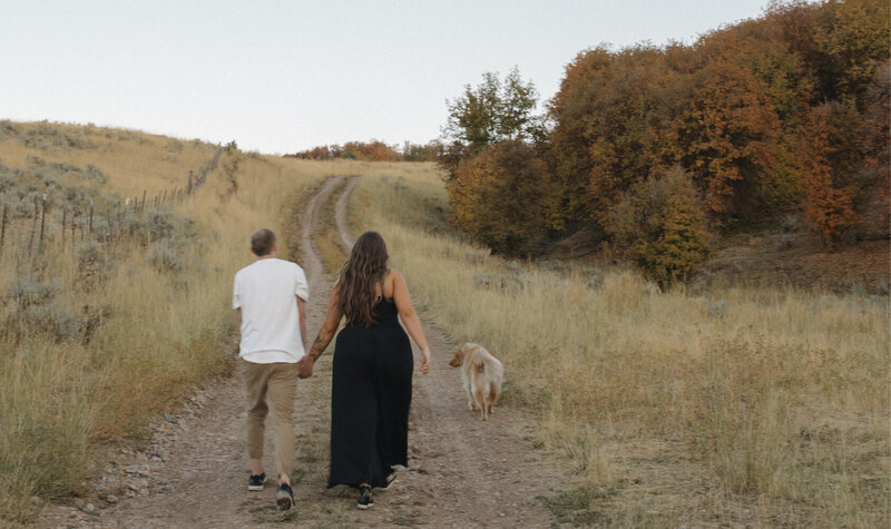 A man and woman holding hands walking away from the camera down a dirt path. Their dog is next to them.