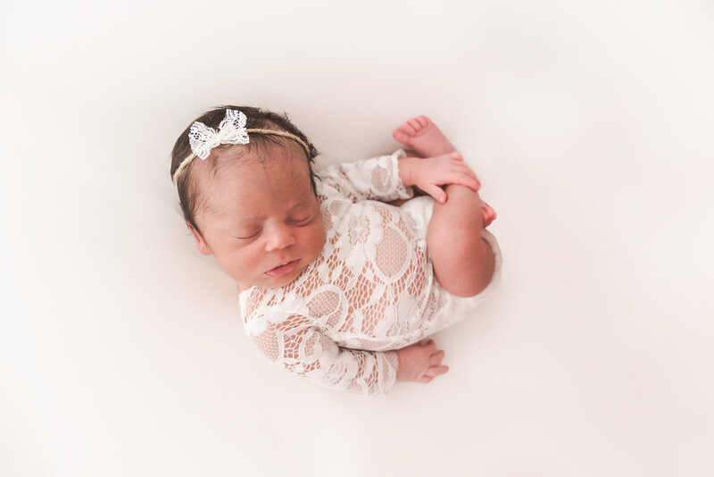 A newborn baby peacefully sleeps on a white background. The baby is wearing a white lace outfit and a delicate headband with a bow. The baby's arms and legs are tucked in close to their body.