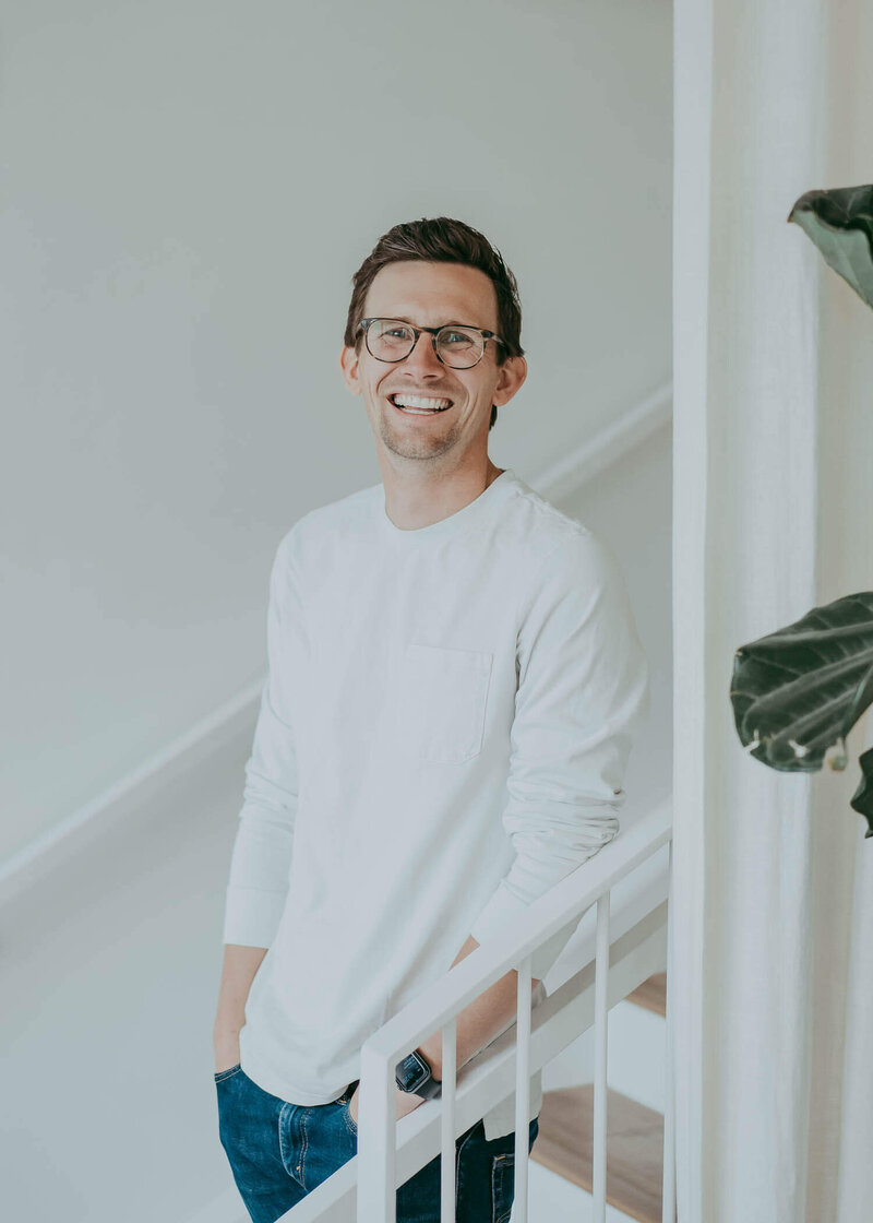 A man wearing a long sleeve white shirt, casually leans against the railing of a staircase during a personal branding session in Denver.