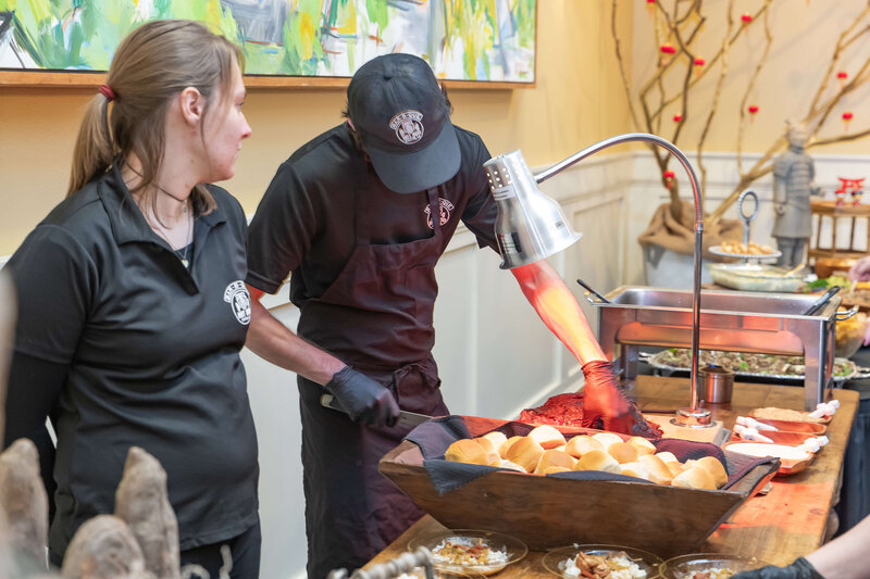 Two catering staff members in black uniforms are preparing food at a buffet table, with one person handling bread rolls under a heat lamp and various dishes arranged around them.